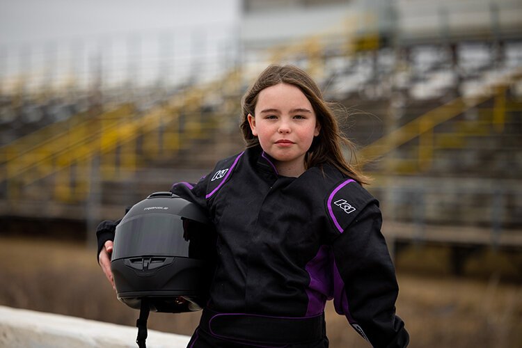 Ella Bringer poses in front of the Mt. Pleasant Speedway's grandstands, wearing her suit and holding her helmet by her side.